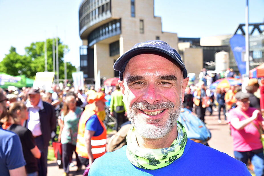Axel Fell, Landesvorsitzender des ADFC NRW Portrait Axel Fell, Landesvorsitzender des ADFC NRW, mit blauem T-Shirt, buntem Halstuch und ADFC-Kappe vor Menschenmenge und Landtag NRW im Hintergrund bei sonnigem Wetter. .